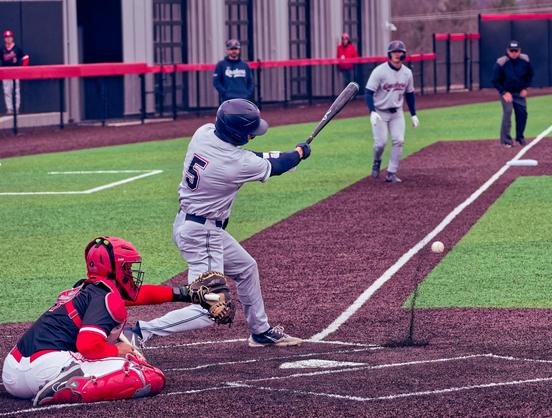 Batter 5 has his bat pointing diagonally up while there is a splash of dirt below the ball that just bounced off the ground, the catcher has his arm out behind him and another Penn player in a grey jersey and blue cap is getting ready to move while a referee in a black jacket looks on