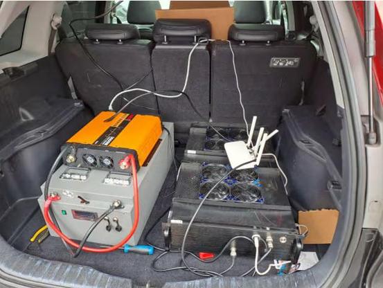 The back of a hatchback where the hatch is up,  several large boxes with many cooling fans are connected with numerous cables including some really huge red and black power leads.