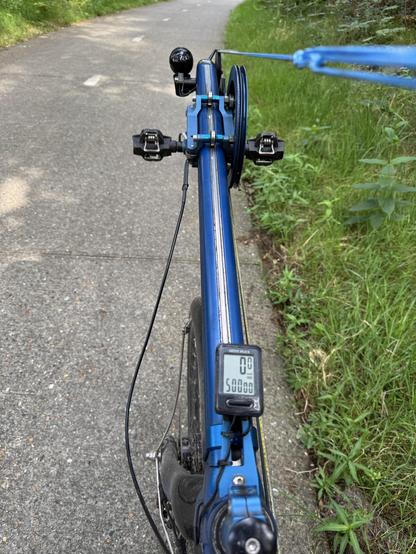 Een betonnen fietspad met ernaast een grasberm. Op het fietspad zie je de boegspriet van een blauwe roeifiets met vooraan een koplamp, dan de revolver ring met aan weerszijden een klikpedaal en op de voorgrond de kilometerteller waarop een totaalstand van 50.000 km staat. 