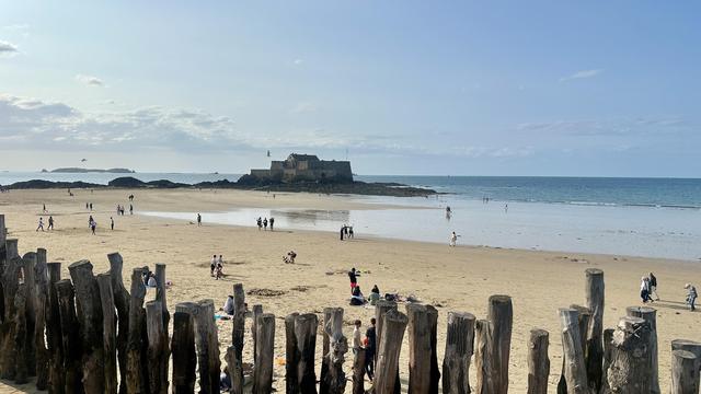 Die Grand-Bé, eine historische Festung auf einer St-Malo vorgelagerten Insel. Aufgrund der Ebbe ist sie gerade trockenen Fusses zu erreichen. Im Vordergrund die bekannten, in den Sand gerammten Baumstämme zum Brechen der Wellen. Menschen flanieren am Strand.