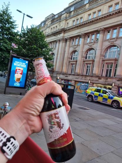 My hand holding a bottle of Saigon beer. In the background a police car and a shopping center.