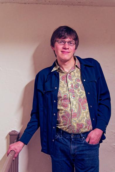 Young man with an almost Beatles haircut and narrow glasses almost smiles while he holds onto a railing with one hand and has the other in his pocket.  He has blue jeans and a blue jacket with 5 buttons on each side and a shirt with a paisley pattern with a definite collar.