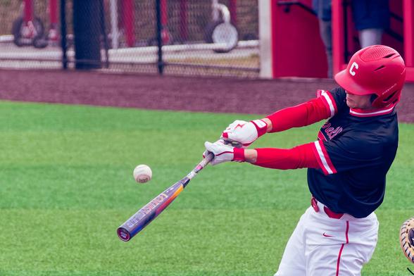 A baseball looks suspended in air above a bat held by a man with white gloves, you can see a little gap of skin and then red sleeves  and a black shirt with a mostly hidden face with a red plastic helmet that has a white C on it and white pants,  over a green synthetic grass field with a brown margin and a fence behind
