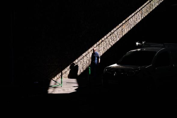 Street photography of a man walking beneath a bridge in Paris, with sunlight shining through the gap between two bridges, illuminating him. A car appears in the shadows behind.