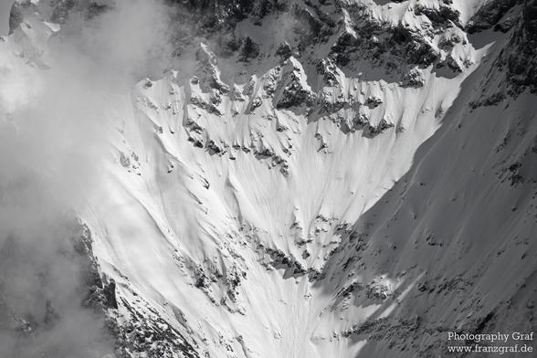 A stunning black and white image of a snow-covered mountain range is captured in this photograph. The mountain is shrouded in clouds, creating a mystical and serene atmosphere. The dominant colors of grey and white add to the peaceful and cold winter scene. The image depicts a glacial landscape with a massif and summit visible in the background. The mountain range is surrounded by fog, giving a sense of mystery and grandeur to the scene. This breathtaking winter landscape is a perfect example o…