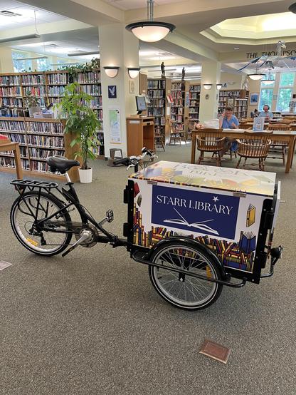 a tricycle with a box on the front for books. It’s a bike-powered bookmobile! in Rhinebeck, NY at the Starr Public Library. In the background you see the usual shelves and tables found in a library. 