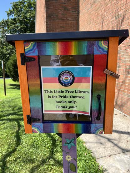 The sign on the front of this Little Free Library says “This little free library is for Pride-themed books only. Thank you.” 
The box is decorated with rainbow colors. 