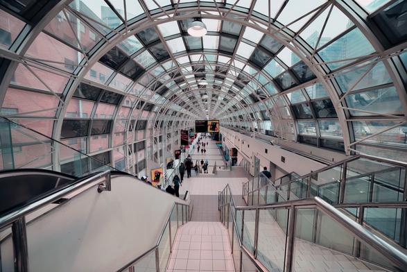 A long, modern enclosed pedestrian skywalk (Toronto Skywalk) with an arched glass and metal ceiling. The tunnel-like corridor stretches into the distance with repeating metal framework creating a pattern overhead. Multiple people walk along the white-tiled floor, some using stairs and escalators in the foreground. Round lights hang from the ceiling at regular intervals. A red brick building is visible through the glass walls on the left side. The perspective is from an elevated viewpoint looking down the length of the walkway.