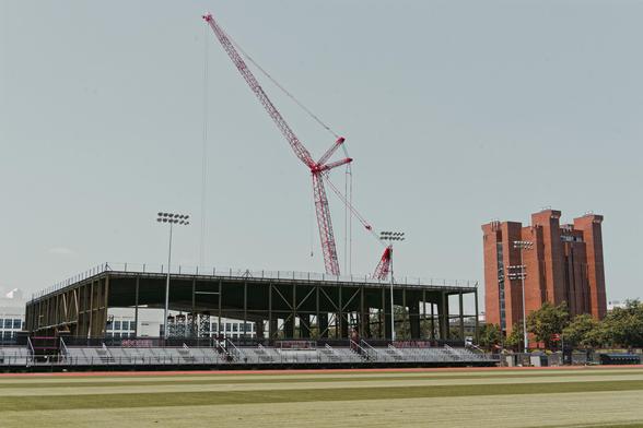 A large crane rises over the metal framework of a building under construction with bleachers and light arrays in the foreground as well as a green athletic field and a ugly brick skyscraper to the far right