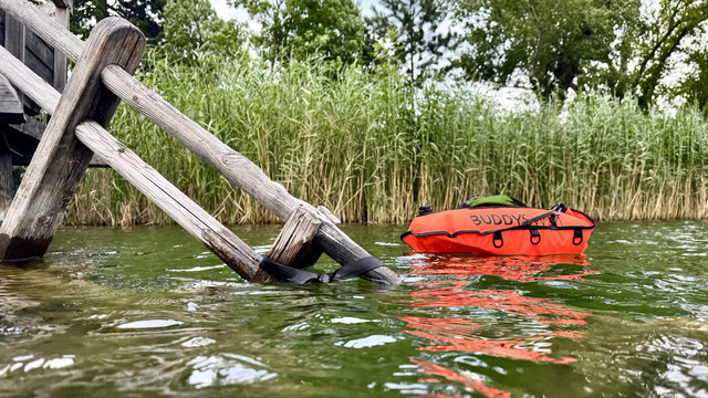 Blick aus meiner Perspektive, wie ich aus dem Wasser steige. Eine schöne alte Holztreppe mit Geländer führt zu einem Steg hinauf. Am Geländer habe ich meine orangefarbene Schwimmboje angebunden. Das Wasser des Sees schimmert grünlich. Im Hintergrund Schilf und Bäume, wolkig-grauer Himmel.