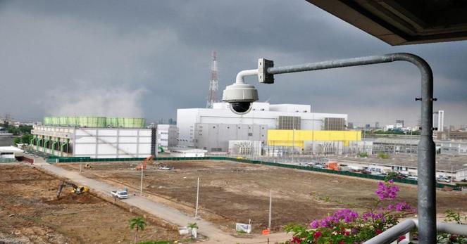 A long shot shows an outdoor CCTV camera on a pole, overlooking a large industrial complex under a cloudy sky. The camera, light gray with a dark lens, extends from a curved metal pole on the right side of the frame. In the background, the industrial complex features several large white buildings, some with green cooling towers emitting steam or vapor. A vast, undeveloped dirt lot with scattered construction equipment and vehicles lies between the camera and the buildings. To the far left, a glimpse of water and what appears to be a port or shipping area is visible. On the right, hints of a city skyline can be seen in the distance. In the foreground, out-of-focus purple flowers add a touch of color to the lower right corner.

