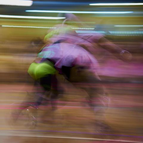 A long exposure photograph of two rollerderby players, one in neon green hotpants and black leotard, the other in a pink tutu. The image is made of motion blurs and horizontal bands of colour, as the camera was panned left to right as the players passed in front.