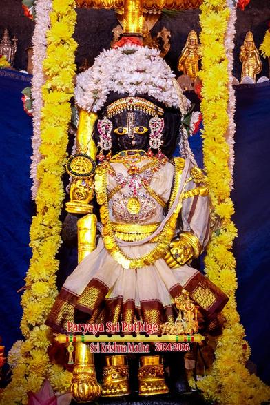 Idol of Hindu deity Krishna in white saree (with red embroidered border) standing with gold-clad staff in right hand and left hand on left front waist; wearing gold shoes, gold palm coverings, and wearing lots of gold and stone-studded ornaments including a large studded medallion of two swans facing each others; and wearing a turban made entirely of white jasmine flowers. Strings of yellow and white flowers fall down on either side of Krishna from a gold umbrella [not visible] above his head.