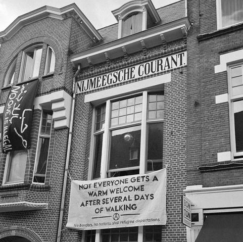 Een zwart-witfoto van een historisch bakstenen gebouw met de naam "Nijmeegsche Courant" op een gevelbord. Aan het gebouw hangen twee spandoeken: links een zwart spandoek met witte tekst in het Arabisch en Engels, waarop staat "No one is illegal"; in het midden een groot wit spandoek met de tekst: "Not everyone gets a warm welcome after several days of walking – No borders, no nations, stop refugee deportations."