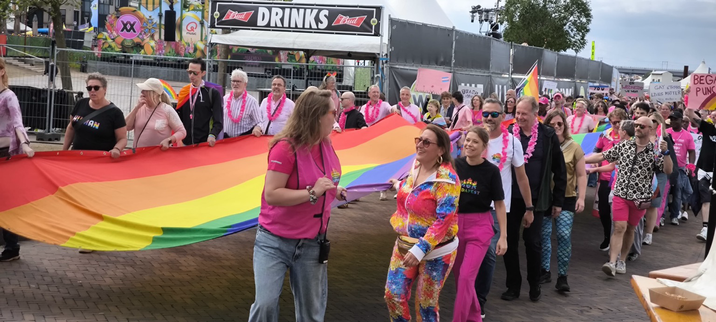 Foto van schuin boven van de optocht. Met een meterslange regenboogvlag tussen de mensen in. Op de achtergrond de rivier De Waal.