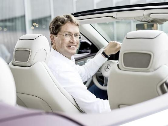 A smiling individual seated in a luxurious Mercedes-Benz convertible, looking towards the camera. The car features a light-colored interior, and the background suggests a modern, upscale environment.