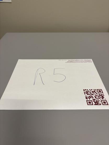 A letter size paper on a desk with a QR code and a caption and the letters R5 handwritten