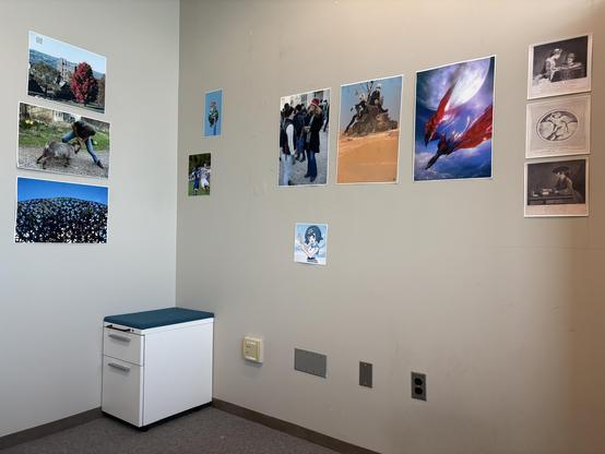 Empty corner in the room with a two-drawer unit with a blue fabric top and many electrical and network outlets and a few rows of small and large prints including family photos, landscapes, video game art and old engravings