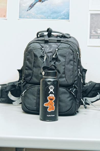 A big ass backpak fills about half the frame,  sitting on a desk that I eliminated with an R5 sigil.  Strapped to the backpack is a big black water bottle with major dents,  a white symbol that looks like an abstract person smiling with hair standing up and a sticker of the Sesame Street character roper in red with a white background and googly eyes and a caption that reads Hydro Flask.  Behind that a few prints are partially visible.
