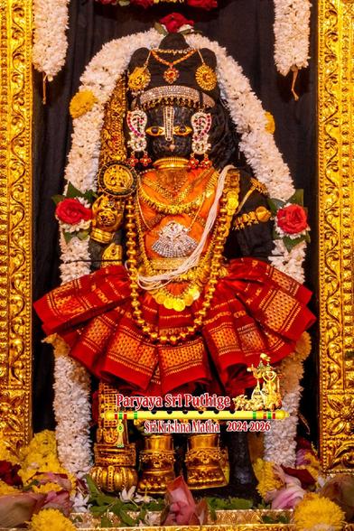 Idol of Hindu deity Krishna in orange vest and red frilled kilt standing on a gold-covered slab with gold-clad staff in right hand; wearing gold shoes and lots of gold ornaments. A thick string of white flowers (with two large red roses) surround Krishna. Just the sides of a gold arch around Krishna are visible.