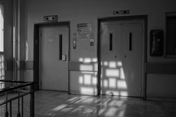 Schwarzweißfotografie:
Zwei unterschiedliche Auzugstüren, dazwischen Hinweisschilder auf griechisch.
Durch ein gekacheltes Fenster fällt ein Muster aus Licht und Schatten auf eine der Türen. Im Vordergrund ist noch ein Stück Geländer zu sehen.
