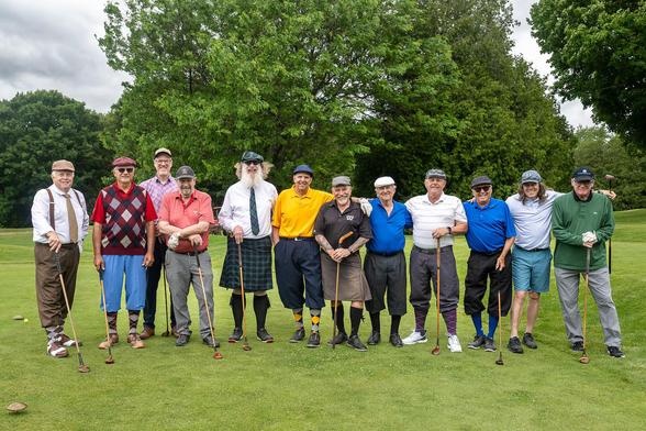 The image shows a group of eleven men standing on a golf course, posing for a photo. They are holding golf clubs and are dressed in a variety of colorful and vintage-style clothing. The men are wearing different types of hats, including flat caps and caps, and some are sporting ties and suspenders. Their attire includes a mix of shorts and long pants, with some wearing knee-length kilts. The men are standing in a line, with some having their arms around each other, suggesting camaraderie. The b…