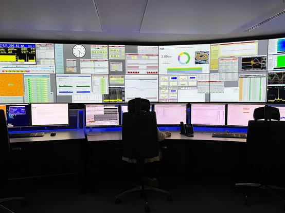 Kommandozentrale, dunkler Raum mit hell erleuchteter großer Monitorwand, auf der allerlei sehr technisch anmutende Daten dargestellt werden.