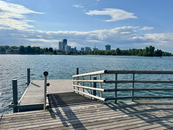 Finally on the shore, where the path continues on foot. View of a beautiful wooden jetty and the water. In the distance, the wooded island of “Gänsehäufel”, a few high-rise buildings, behind them the range of hills of the Vienna Woods. Blue sky, low cloud cover in the distance.