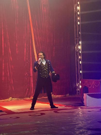 A performer dressed in a historical-style costume stands on a stage, holding a microphone with one hand and a hat in the other. The background features red velvet curtains and stage lights, creating a theatrical atmosphere.