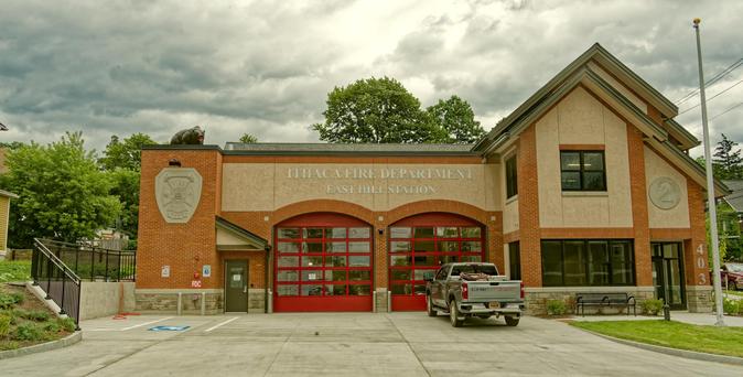 A firehouse with two red doors with lots of windows and a pickup truck in front as well as two colors of brown brick and a complex roofline on the side,  not to mention a flagpole without a flag.