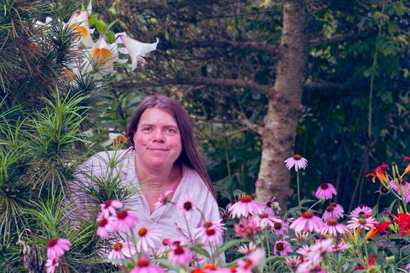 Woman sits in a garden with an echinacea field in front,  lily flowers behind and surrounded by various evergeen trees