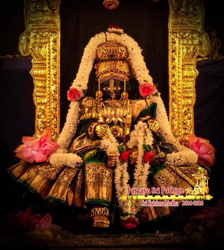 Idol of Hindu deity Krishna in green saree (with a broad embroidered border) seated with right leg hanging down, left leg folded at the knee  resting on the seat, and left foot touching the inside of the right knee. Krishna wears gold shoes, gold palm coverings, gold crown, and several gold jewels including a huge lotus-shaped gold medallion. Krishna holds a garland of white jasmine flowers adorned with three large red roses. Two huge pink lotus blooms on either side of Krishna. The saree is pleated and the pleats are spread in a radiant circular manner to cover Krishna's legs with just the right shoe peeping out.

A thick string of white flowers go from the left side of Krishna, over his crown, and back down to Krishna's right side. The sides of a gold arch behind Krishna are visible. Dark almost-black background.