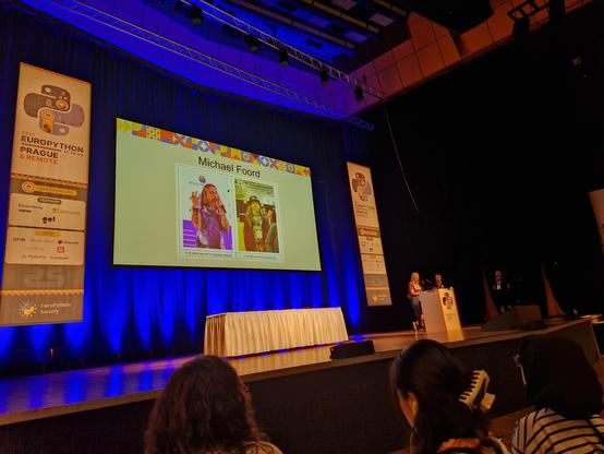Slide at EuroPython main stage with two photos of Michael Foord in colourful clothes, one (singing?) with a mic, the other wearing a top hat with steampunk goggles on the brim. 
