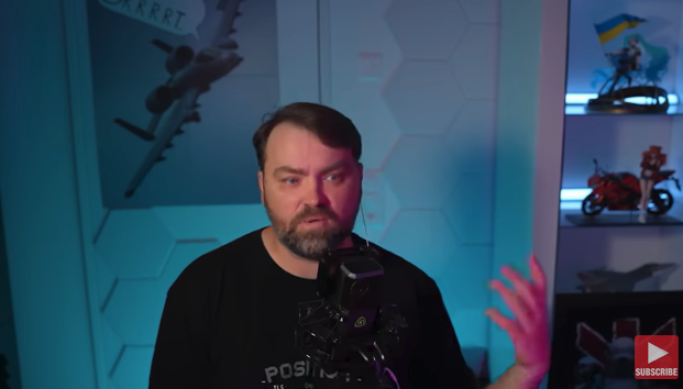 A man with short hair that makes a sinusoidal curve across his forehead has a microphone in front of him an a picture of an A-10 Warthog behind him and his hand stretched out with his palm facing back at him.  At the far left of the frame is a set of cubbyholes,  the topmost of which has a girl with long turquoise twintails waving a Ukraine flag,  below that is a girl with red hair an an open midriff standing besides a red motorcycle.