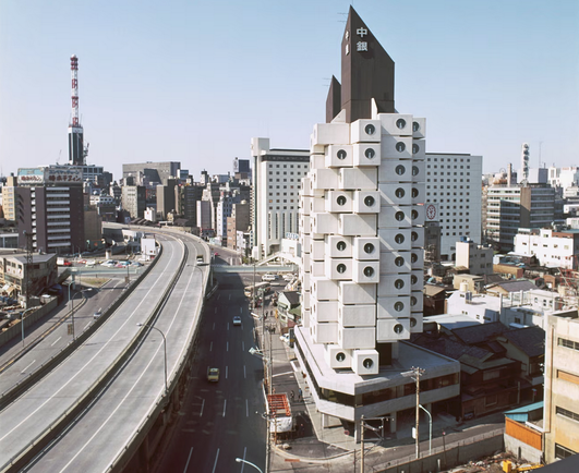 A bit of left of center: A skyscraper with two brown cores that come to a point at the top are surrounded by a number of white blocks with circular windows that have a line in the bottom.  Multi-leveled roads run back to front in the mid-right of the frame while other buildings fill out the city.