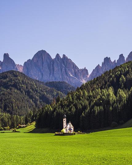 Church of St. John / Chiesetta di San Giovanni in Ranui, Funes, Italy, nestled in a vibrant green meadow with dense forest and dramatic mountain peaks in the background. The quaint church features a small bell tower with a dome, set against a clear blue sky in a serene alpine landscape.