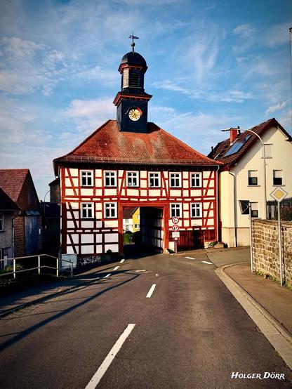 Blick aus Südwest: Frontalaufnahme eines barocken Fachwerkhauses mit Uhrturm in Lich-Oberbessingen. Das rot-weiß gestrichene Torhaus besitzt eine große Durchfahrt, durch die eine Landstraße führt. Die warme Abendsonne taucht Dach und Fassade in goldenes Licht. Links und rechts schließen sich Dorfhäuser an.