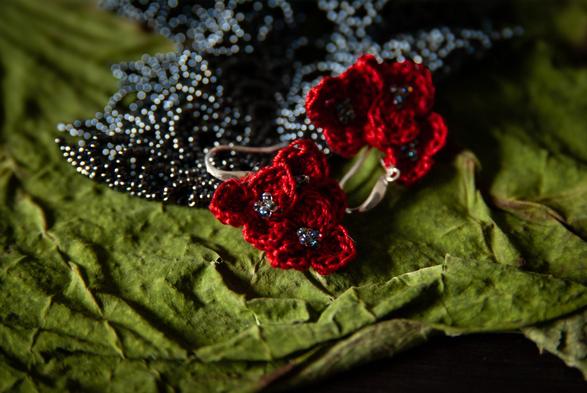"Close-up of handmade crochet earrings featuring deep red thread flowers with small glass bead centers, resting on textured green leaves and a background of dark, shimmering beads."