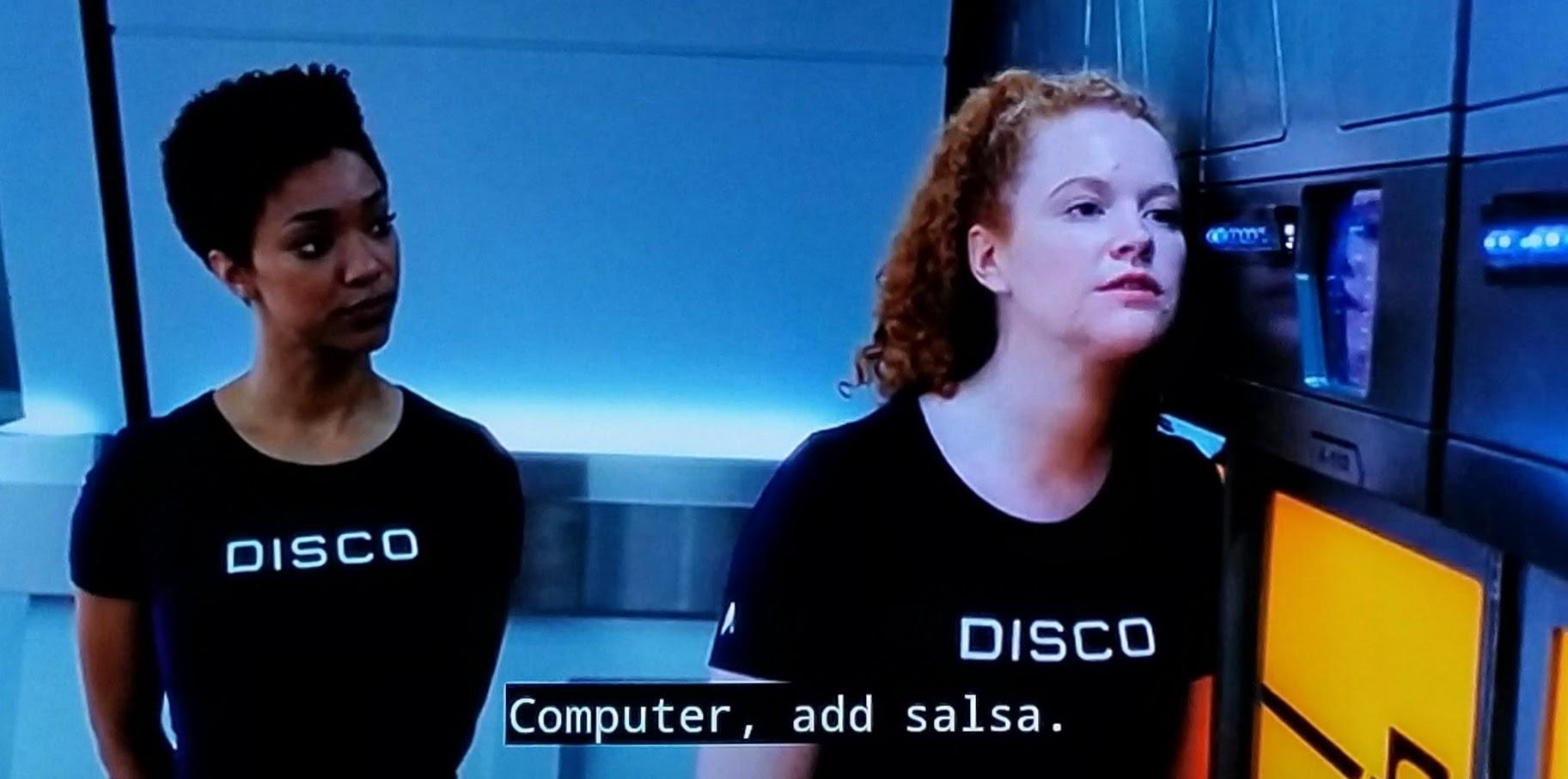 Star Trek Discovery scene. Michael Burnham and Tilly are in the dining hall. Both are wearing gym clothes that say DISCO on the front. Burnham is standing and observing Tilly like a disappointed teacher. Tilly is leaning in towards a food replicator, extending her head towards its panel. Closed caption reads, "Computer, add salsa."