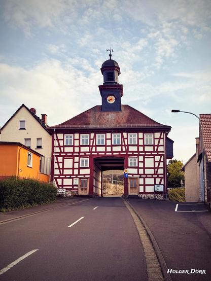 Blick aus Nord-Ost: Frontalansicht eines historischen Torhauses mit markantem Fachwerk und Uhrturm in Oberbessingen, aufgenommen von der Straße aus. Der Blick führt durch die zentrale Durchfahrt hindurch bis zur rückseitigen Mauer. Links und rechts rahmen Wohnhäuser das Bild ein, der Himmel ist teils bewölkt.