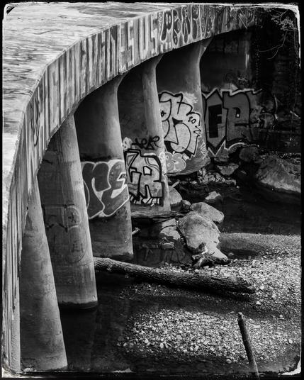 Black and white photo of the graffiti covered supporting pillars of a curved footbridge over a small river.
