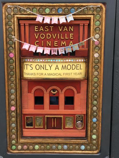 The image features a decorative sign that resembles a miniature cinema facade. The sign is framed in gold with colorful, circular embellishments along the border. At the top, the text "EAST VAN VODVILLE CINEMA" is prominently displayed in gold letters on a red background. Below this, a banner reads "HAPPY BIRTHDAY!" in pastel-colored letters. The central part of the sign includes a yellow banner with the text "IT'S ONLY A MODEL" and "THANKS FOR A MAGICAL FIRST YEAR!" in black letters. The lower…