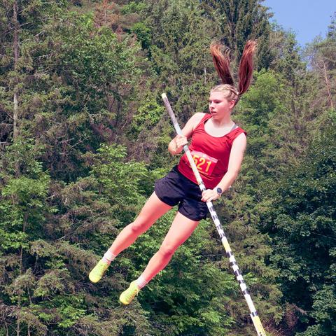A woman wearing a red tank top, green shoes and black shorts holds a vaulting pole as she flies through the hair with two twintails up over her head