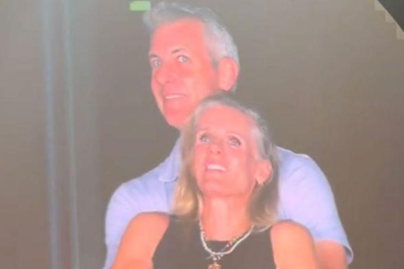 The image shows two individuals in a dimly lit environment. The person in the foreground is wearing a black sleeveless top and a necklace with a pendant. The person in the background is wearing a light blue short-sleeved shirt. The lighting in the image is low, creating a hazy effect, and the background is indistinct, with a faint outline of a staircase visible on the right side. The individuals appear to be in close proximity, with the person in the background slightly behind the person in the…