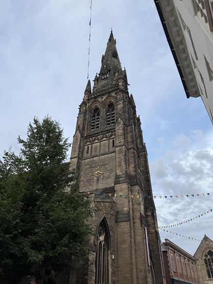 A tall, historic stone church tower rises against a gray sky, adorned with decorative bunting. Surrounding trees and nearby buildings create a quaint street scene.