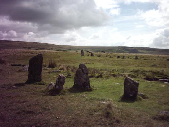A colour photo looking across the eastern ends of both the, roughly parallel, double stone rows of small standing stones at Merrivale on Dartmoor in Devon. Four stones of the northern avenue are nearest the camera with a greater number of the southern avenue in the middle distance. The undulating moors stretch into the distance and the sky is full of grey and white clouds, with small areas of blue showing.