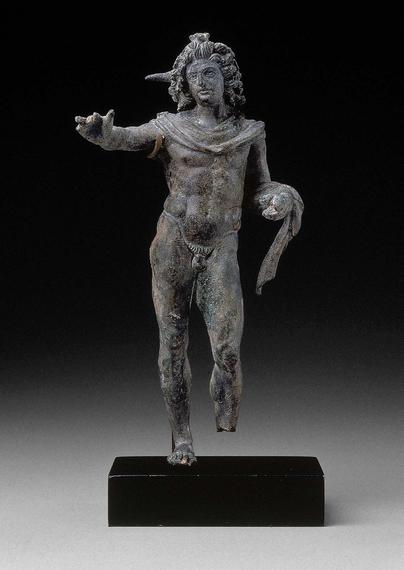 Bronze statuette of Helios, the sun god. The sun god is young and beardless but powerful, with a lion-like mane of hair. Like Alexander, he was imagined as fast-moving and far-seeing. Beams of light, of which only one remains, originally radiated from his head. Helios travelled through the heavens in a chariot, and he probably once held a whip in his left hand around which also drapes his himation. His right arm is raised, maybe holding the reins of his chariot.