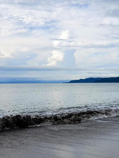 A wave breaks on a dark sand beach in Drake Bay, on the Pacific coast of Costa Rica. On the horizon is a darkly forested headland. A towering cumulo-nimbus cloud rises into the sky in the distance 