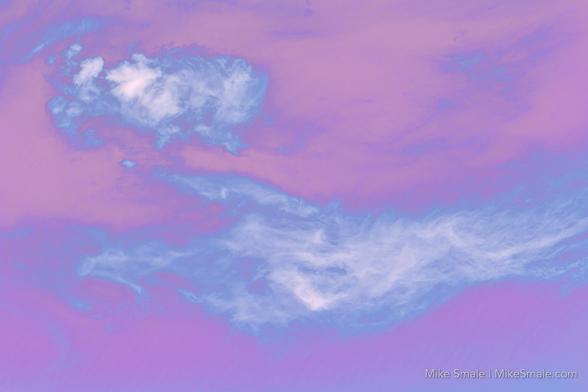 Delicate pastel hues blend to create a dreamy skyscape with soft clouds. Wispy formations float against a purple and pink background, evoking a serene atmosphere.
