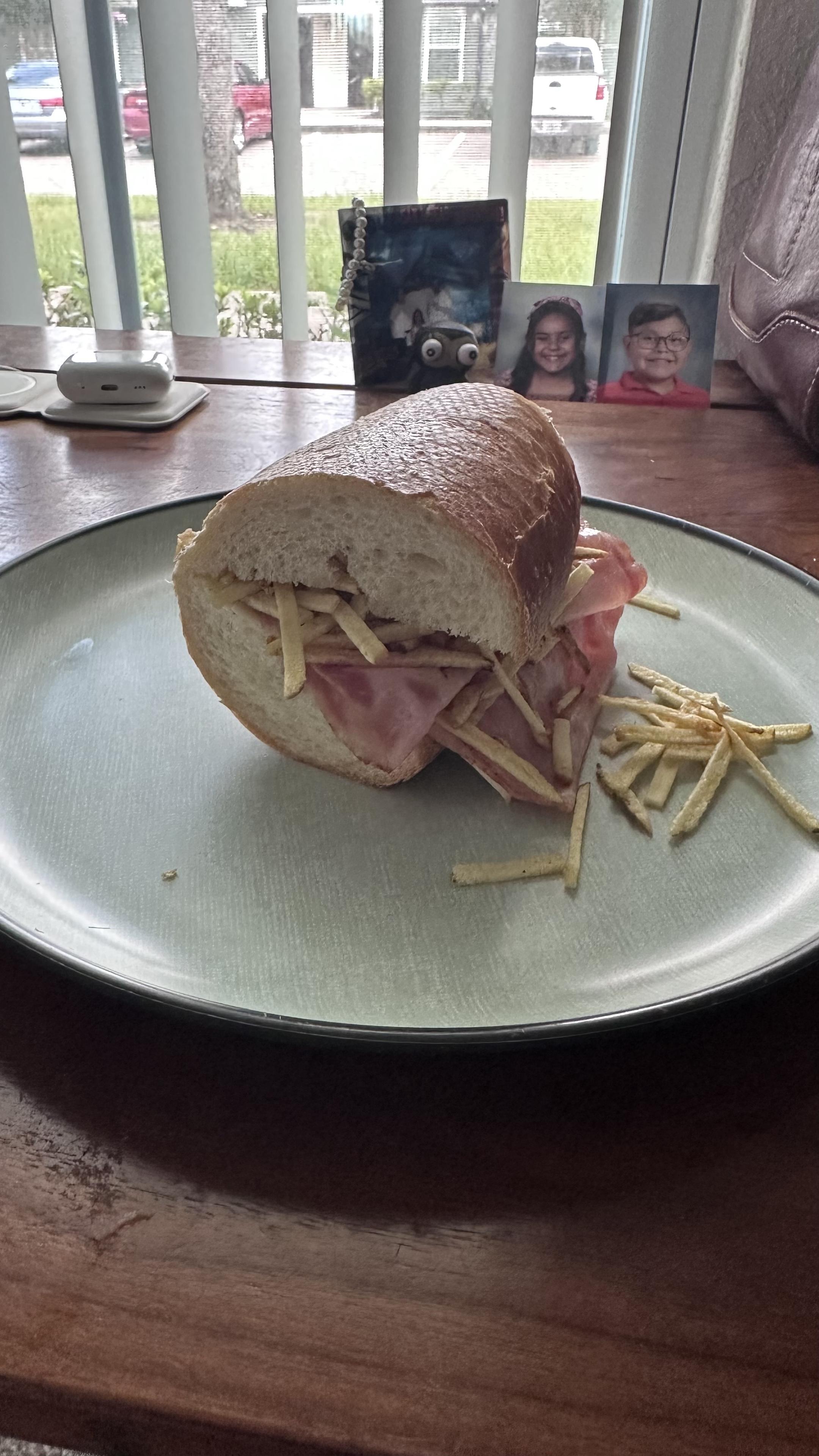 A sandwich is placed on a green plate, featuring ham and string potatoe chips inside a partially opened bread roll. In the background, there are framed photos and a small decorative item on a wooden table.
